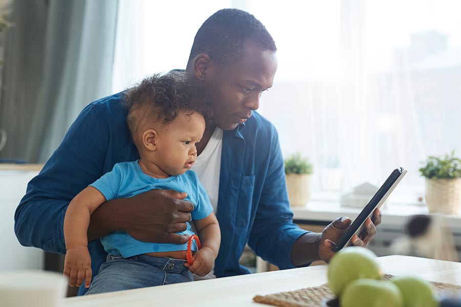 Client Center - Father Uses Laptop and Phone at Kitchen Table With His Toddler Son on His Lap Playing With a Ring Toy